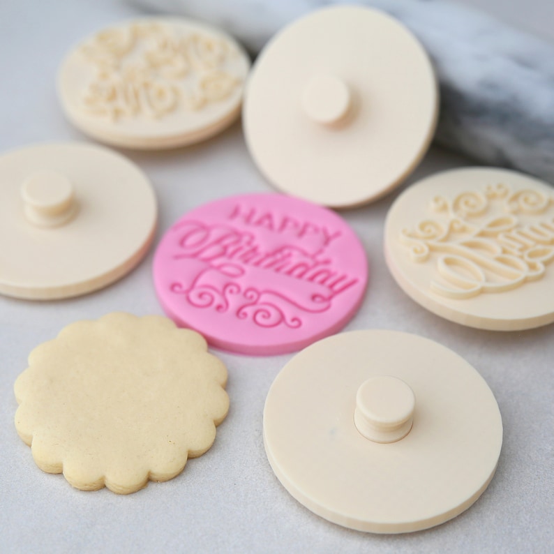 May include: A set of six cookie stamps, including four plain white stamps and two decorated stamps. One decorated stamp is pink and says "Happy Birthday" in a cursive font. The other decorated stamp is white and has a cursive design. A plain cookie is also shown in the image.