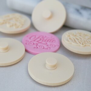 May include: Five round, white, plastic cake decorating stamps with a knob on top. One stamp is pink and has the word "Bride" written in cursive.