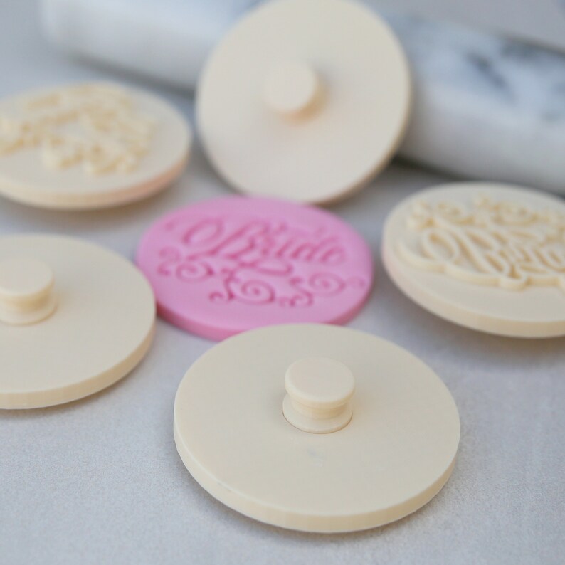 May include: Five round, white, plastic cake decorating stamps with a knob on top. One stamp is pink and has the word "Bride" written in cursive.
