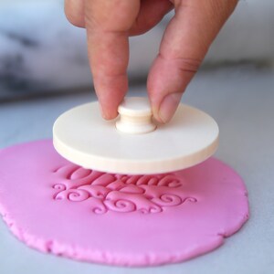 May include: A white plastic stamp with a wooden handle is being pressed into pink fondant. The stamp creates a design of swirls and the word "Welcome".