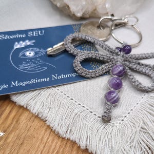 Peut inclure: Un porte-clés en macramé gris avec trois perles violettes et blanches rayées. Le porte-clés est à côté d'une carte de visite avec le texte "Séverine SEU Iridologie Magnétisme Naturopathe". Un jeu de clés est attaché au porte-clés.