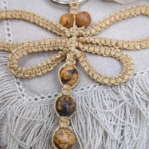 Peut inclure: Un porte-clés macramé fait à la main avec un motif de papillon. Le porte-clés est fait de corde beige et comporte trois perles décoratives mouchetées marron et jaune. Le porte-clés est attaché à un anneau en métal argenté.