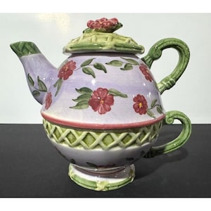May include: A decorative ceramic teapot with a lavender body, decorated with red floral patterns and green leaves. The teapot has a green handle and spout, and a matching lid with a floral accent. The base has a green and red trim.