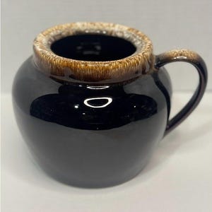 May include: A black ceramic mug with a rounded body and a handle. The rim and handle are brown, and the interior is dark. The mug has a glossy finish and appears to be handmade.