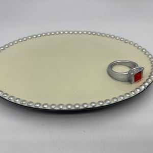Puede incluir: Un plato para anillos blanco con un borde de cuentas plateadas y un anillo de plata con una piedra preciosa roja.