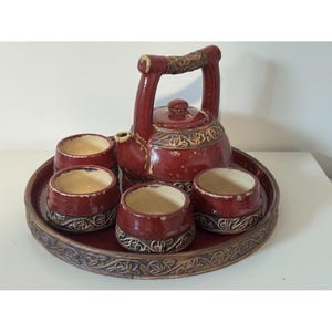 May include: A red ceramic tea set with a teapot, four cups, and a tray. The teapot has a handle and lid, and the cups and teapot have a decorative pattern. The tray has a raised edge with a carved design. The set is on a white surface.