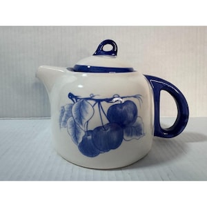 May include: A white ceramic teapot with a blue cherry design. The teapot has a curved spout, a rounded body, and a blue handle. The lid is also white with a blue trim and a small loop handle.