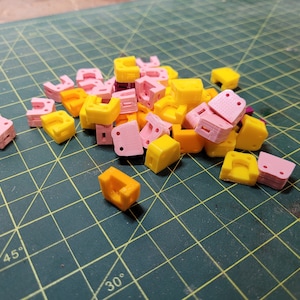 Peut inclure: Un tas de petits supports d'angle en plastique imprimés en 3D, de couleur rose et jaune. Les supports sont disposés de manière aléatoire sur un tapis de coupe vert.