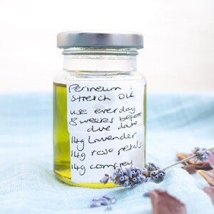 May include: A clear glass jar with a silver lid filled with a yellow liquid. The jar has a handwritten label that reads "Perineum Stretch Out use everyday 8 weeks before due date 14g Lavender 14g rose petals 14g comfrey".