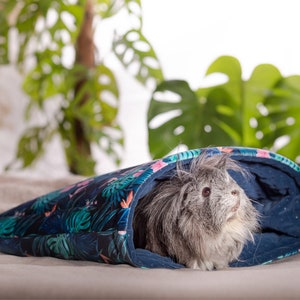 May include: A gray and white guinea pig is resting inside a blue and green patterned fabric sleeping bag. The sleeping bag is on a light brown surface.