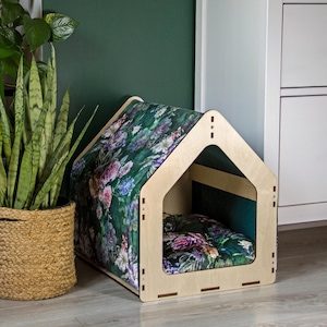May include: A wooden dog house with a green floral patterned cushion inside. The house is light brown and has a simple design.