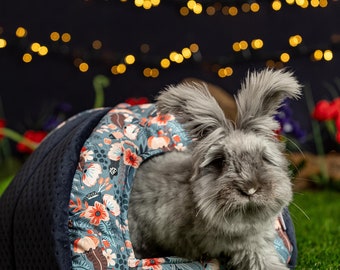 Peut inclure: Un lapin angora gris à poils longs est assis dans un lit bleu à motifs floraux. Le lit est en forme de tunnel et a une ouverture ronde. Le lapin regarde la caméra.