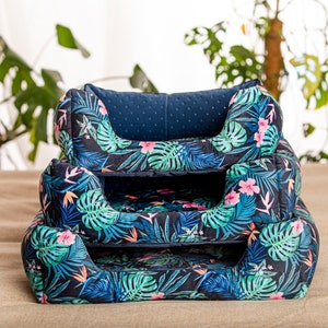 Peut inclure: Un ensemble de trois lits pour chien de différentes tailles, avec un imprimé de feuilles tropicales et des fleurs roses sur un fond bleu foncé. Les lits sont faits d'un tissu doux et confortable et ont un bord surélevé pour un soutien supplémentaire.