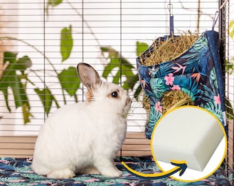 May include: A white rabbit in a cage with a hanging hay feeder. The feeder is made of fabric with a tropical print and is filled with hay. The rabbit is looking at the feeder.