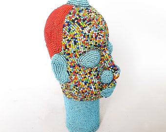 May include: A beaded sculpture of a human head with a blue, red, and multi-colored pattern. The head is on a blue beaded stand.