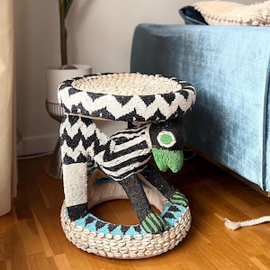 May include: A black and white beaded stool with a green and blue beaded animal figure. The stool is round and has a woven top.