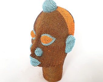 May include: A beaded sculpture of a human head with a blue and orange design. The head is brown and has a blue beaded crown.