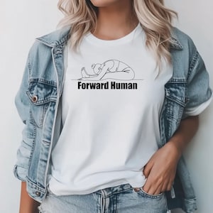 Puede incluir: Camiseta blanca con un dibujo lineal negro de una persona en una postura de yoga de flexión hacia adelante. El texto "Forward Human" está impreso en negro debajo de la ilustración. La modelo lleva una chaqueta vaquera.