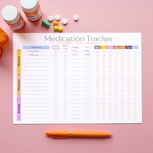 Op de afbeelding: Een medicatietracker met de titel "Medication Tracker" wordt weergegeven op een roze oppervlak. De tracker bevat secties voor medicatie, dosering en tijd, met selectievakjes voor elke dag van de week. Een oranje pen ligt onderaan.