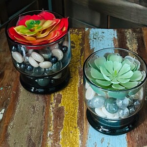 May include: Two glass containers filled with smooth, polished stones and artificial succulents. One container holds a vibrant red and yellow succulent, while the other features a light green succulent. The stones are a mix of white, gray, and black.