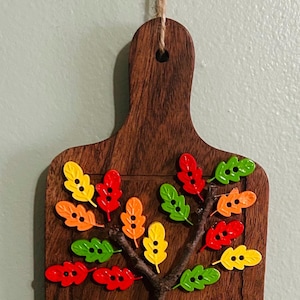May include: A rectangular wooden cutting board with a handle and a jute twine hanger. A small tree branch is attached to the center, decorated with numerous colorful leaf-shaped buttons in shades of red, orange, yellow, and green.
