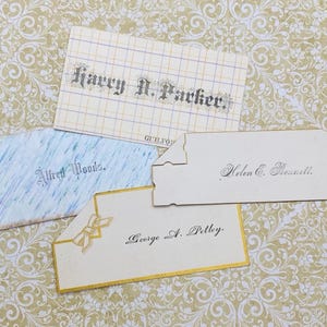 May include: Four vintage place cards with handwritten names in cursive script. The cards are white with gold trim and feature different designs. The names on the cards are Harry N. Packer, Alfred Woods, Helen C. Bennett, and George A. Petley.