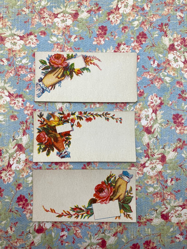 May include: Three vintage blank cards with floral designs. Each card features a hand holding a blank card with a floral design. The cards are on a blue and white floral background.