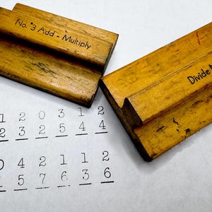 May include: Two vintage wooden math tools with text. One reads "No. 3 Add - Multiply" and the other "Divide No. 4." Below are printed math problems with numbers. The tools are a warm, aged wood color.