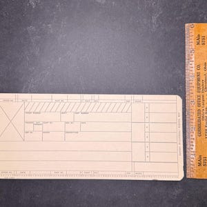 May include: A rectangular tan paper job ticket form with black printed grid lines and text, including fields for order number, date, and part number. A wooden ruler placed vertically next to the form shows a length of approximately 6 inches.