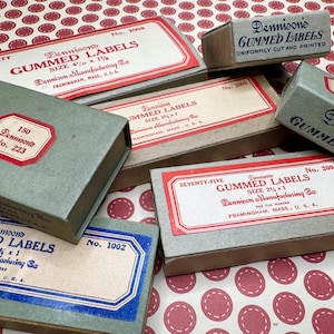 Op de afbeelding: Een verzameling vintage Dennison's gummed labels in verschillende maten en hoeveelheden. De rechthoekige dozen hebben rode en blauwe accenten en bevatten tekst zoals "GUMMED LABELS" en "FRAMINGHAM, MASS., U.S.A."