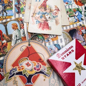 May include: A collection of vintage paper ephemera including a card with the letter B and children, a round hanging ornament with a floral basket illustration, and a red and white box labeled Dennison Stars with a gold foil star graphic.