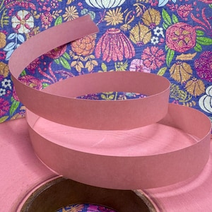 May include: A roll of pink paper ribbon with a spiraling section, set against a floral patterned background in shades of blue, pink, and gold. The ribbon is a solid, muted pink color.