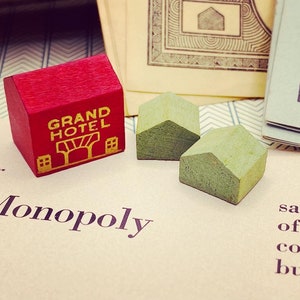 Könnte beinhalten: Ein rotes, hölzernes Monopoly-Spielstück mit der Aufschrift "Grand Hotel" in goldenen Buchstaben. Es gibt auch drei kleinere grüne Holzhäuser.