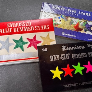 Op de afbeelding: Drie vintage dozen Dennison gummi-sterren. Eén doos leest "Embossed Metallic Gummed Stars" met blauwe, zilveren, groene, rode en gouden sterren. Een andere doos zegt "Forty-Five Stars Assorted". De derde doos zegt "Day-Glo Gummed Stars".