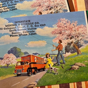 May include: Vintage postcard featuring a moving truck with the text "ALLIED" and a family scene. The illustration shows a truck on a road, a man, a girl in a yellow dress, and a dog. Cherry blossom trees and text for Interstate Moving & Storage Co.