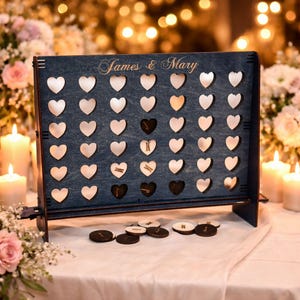 May include: A dark blue wooden game board featuring a grid of heart-shaped cutouts and the engraved text James & Mary. The board includes several circular wooden game pieces with letters scattered at the base, designed for a connect-style game.