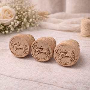 Three wooden cork stoppers with engraved names and dates, placed on a white surface with a floral arrangement in the background.