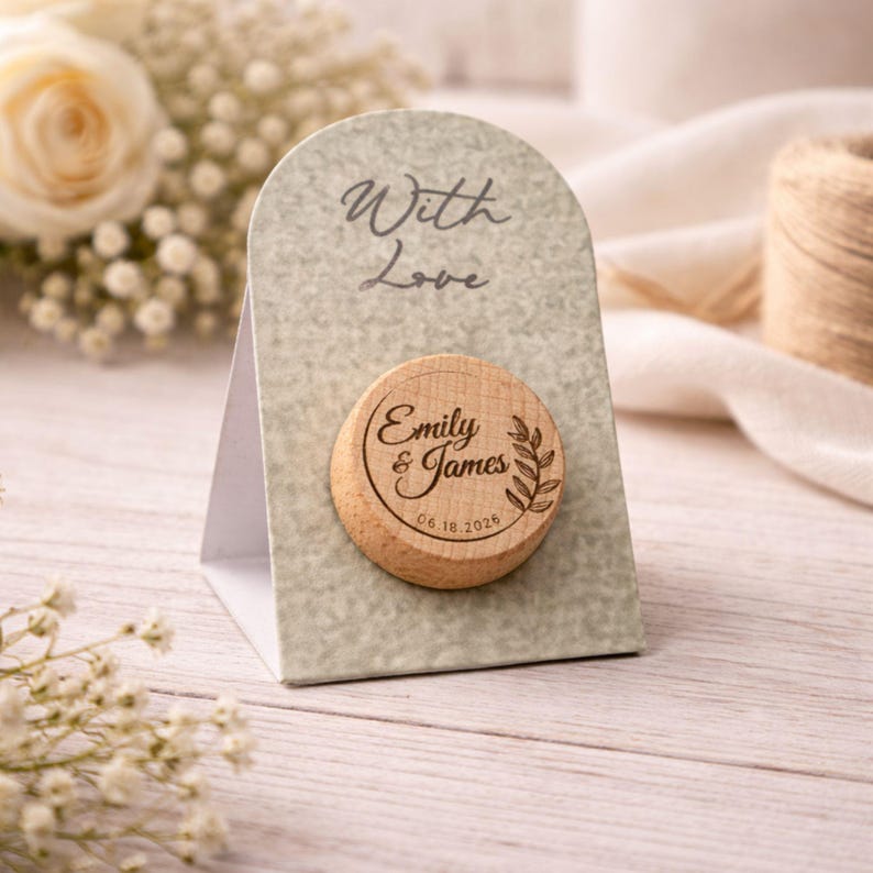 A wooden coaster with a round magnet attached to it, displaying the names 'Emma & James' and the date '06.16.2016'. The coaster is placed on a wooden surface, with a bouquet of white flowers in the background.
