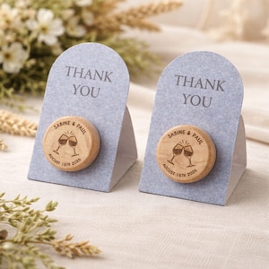 Può includere: Due tappi per bottiglia in legno con un design a calice e i nomi "Sabine & Paul" e la data "18 agosto 2026" incisi. Ogni tappo è esposto su un biglietto grigio con la scritta "THANK YOU".