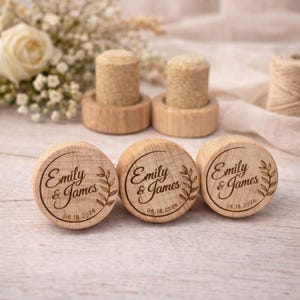 Three wooden cork stoppers with engraved names and dates, placed on a wooden surface with a floral arrangement in the background.