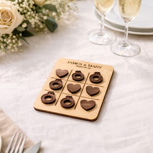 A wooden tic-tac-toe board with engraved rings and hearts on it, placed on a table with champagne glasses and a floral arrangement.