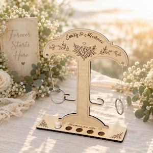 Op de afbeelding: Een houten ringwerpspel met de namen "Emily & Michael" gegraveerd bovenaan en de zin "Hooked for Life" onderaan. Het spel heeft een T-vormig ontwerp met bloemmotieven en bevat metalen ringen.