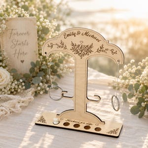 Op de afbeelding: Een lichtgekleurde houten ringwerpspelstandaard met de gegraveerde namen Emily & Michael, bloemillustraties en de tekst Hooked for Life. De standaard bevat twee hangende metalen ringen en een voet met vier ronde uitsparingen.