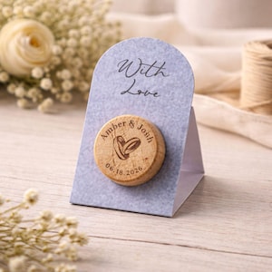 A gray paper stand with a wooden coaster-shaped magnet on it, displaying the text "With Love" and the names "Amber & Josh" along with the date "06.18.2026".