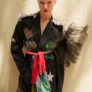 Può includere: Blazer nero con dettagli patchwork, una fascia rossa e un accento in tulle nero. Il blazer presenta una toppa a forma di cuore, una stella e vari ritagli di tessuto. L'outfit è completato da leggings fantasia.
