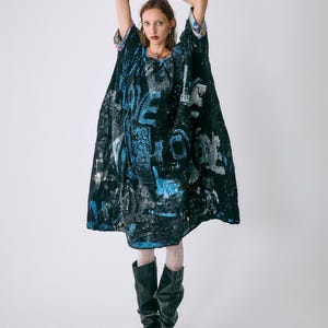 May include: A black, blue, and white patterned dress with a loose fit. The dress features a unique design with text and abstract shapes. The model is wearing black boots and white stockings. The sleeves have colorful patterned trim.