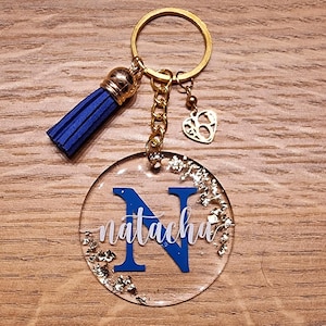 Peut inclure: Un porte-clés bleu et or avec un charme rond en résine représentant la lettre "N" et le nom "natacha" en lettres blanches. Le charme est rempli de paillettes dorées. Le porte-clés a également un pompon bleu et un charme en forme de cœur doré.