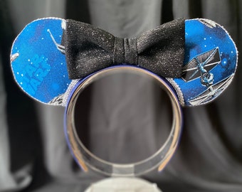 Puede incluir: Orejas de Minnie Mouse de color azul y blanco con temática de Star Wars con un lazo negro brillante. Las orejas presentan una nave espacial y el texto "Star Wars".