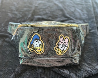 May include: Black fanny pack with a gold zipper and glitter. It features embroidered patches of Donald Duck and Daisy Duck.
