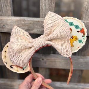 May include: A pair of beige burlap mouse ears with a large beige burlap bow. The ears are decorated with a white fabric featuring a geometric pattern and a cartoon lion.